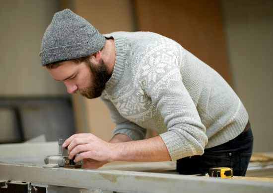 Ein junger Mann mit grauer Wollmütze und hellgrauen Weihnachtspullover steht gebeugt über einer Holzleiste und arbeitet daran.