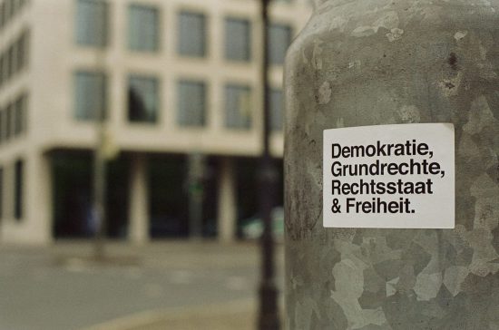 Auf einer Säule stehen die Worte "Demokratie, Grundrechte, Rechtsstaat & Freiheit." Im Hintergrund sieht man in der Ferne ein Gebäude mit vielen Glasfenstern.